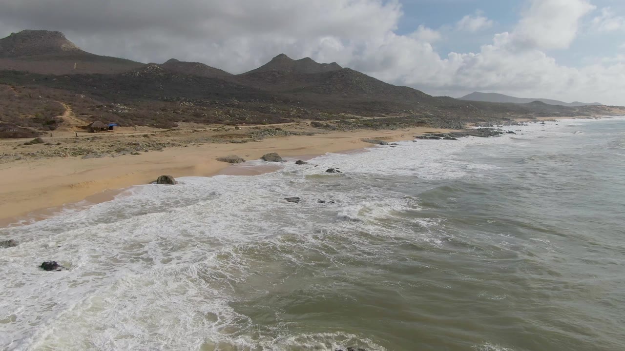 At 0:03, the surf spreads across a broader section of Punta Gorda beach as rocks interrupt the foam and dark mountains sit beneath cloud shadow, with no visible people.