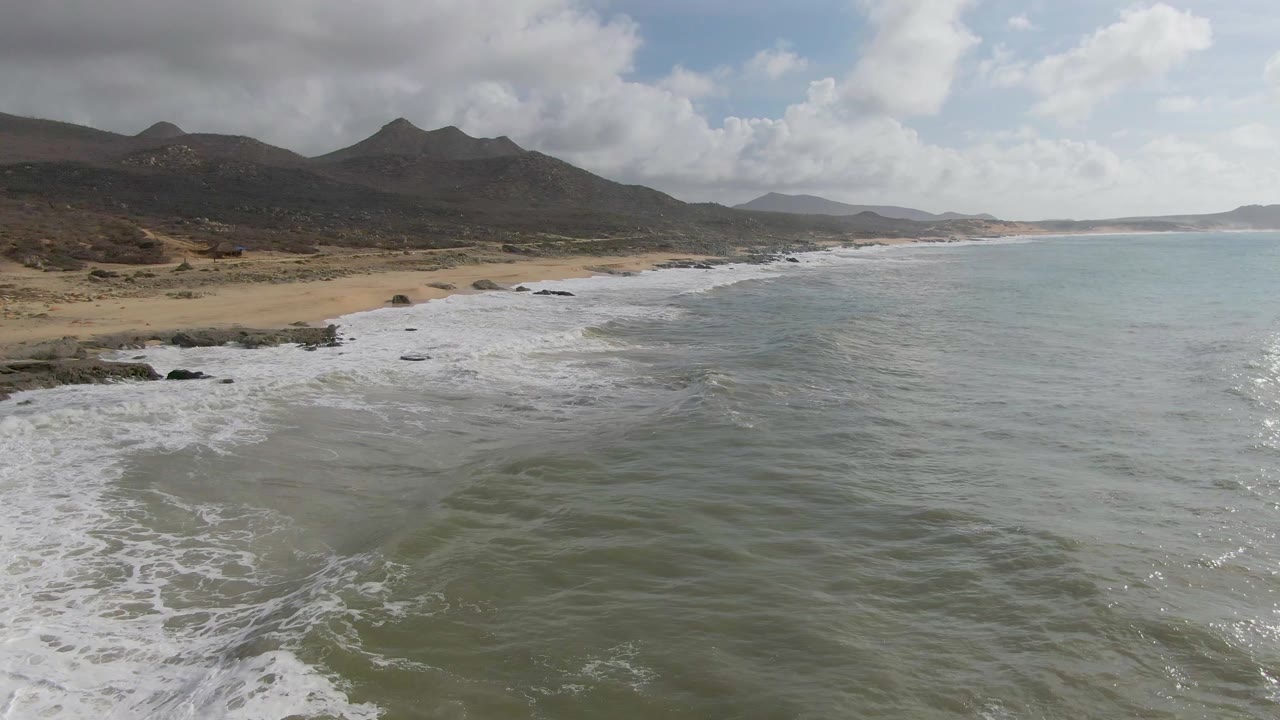 Elevated view of Punta Gorda East Cape surf rolling toward tan sand, with dark rocky outcrops, low mountains, partly cloudy sky, and no visible people.