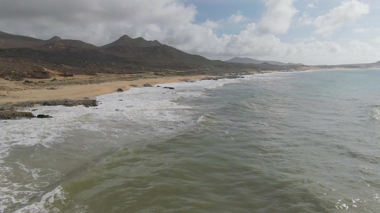 Punta Gorda East Cape shoreline from above, with more white foam across the near shallows, tan beach at left, dark mountain ridges inland, and no visible people.