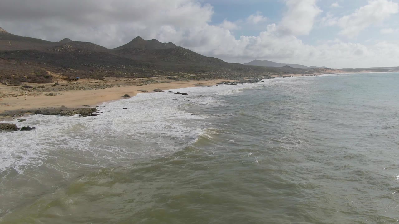 Aerial Punta Gorda East Cape view with broken whitewater near rocky points, open green-gray sea to the right, tan sand to the left, mountains inland, and no visible people.