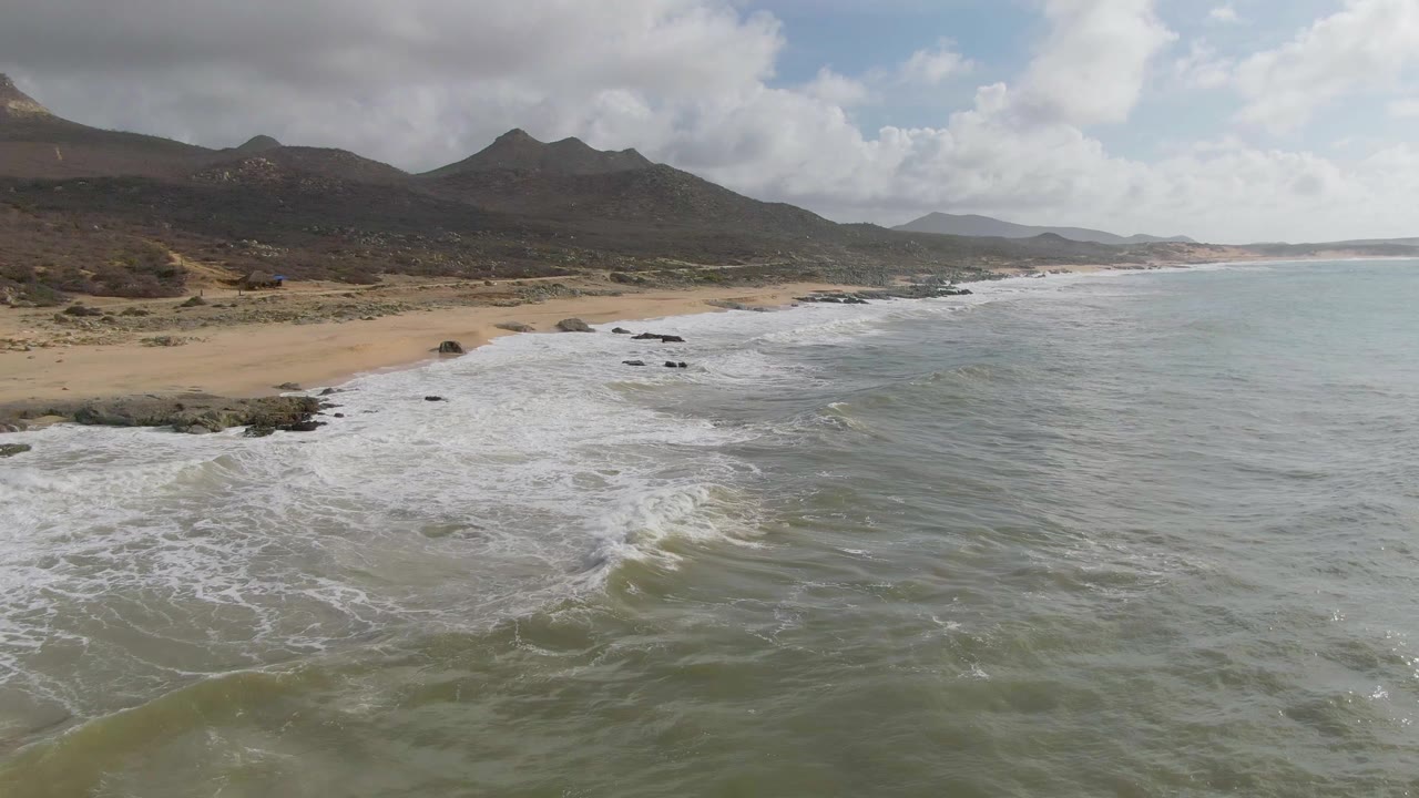 Surf washes toward sand with rocky shoreline and mountains in the distance