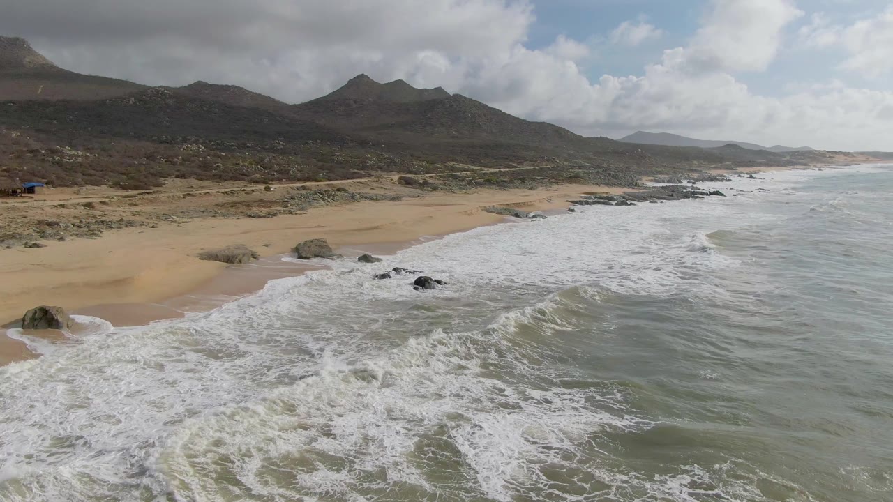 Aerial view at Punta Gorda, Baja California Sur, showing white surf crossing dark rocks onto tan beach with mountains and clouds in the background, no visible people.