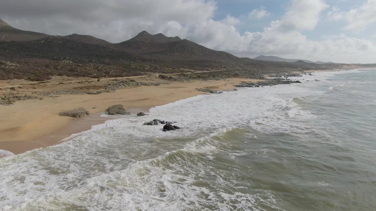 At 0:01, foamy ocean water spreads farther across the rocks and beach at Punta Gorda, with the mountain ridge still visible under broken cloud, no visible people.