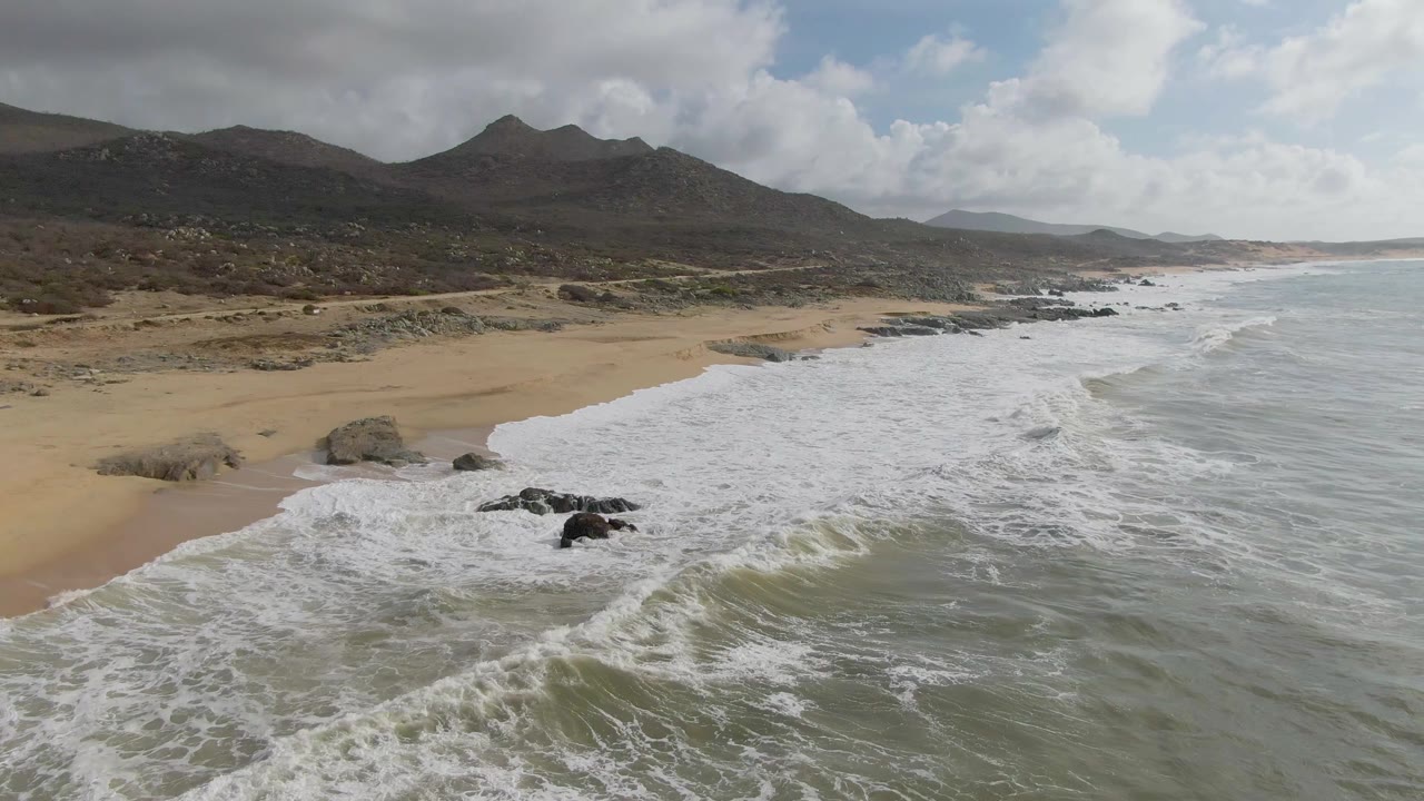 At 0:02, a broad sheet of white surf advances along the sandy Punta Gorda shoreline, with black rocks in the water and low mountains behind, no visible people.