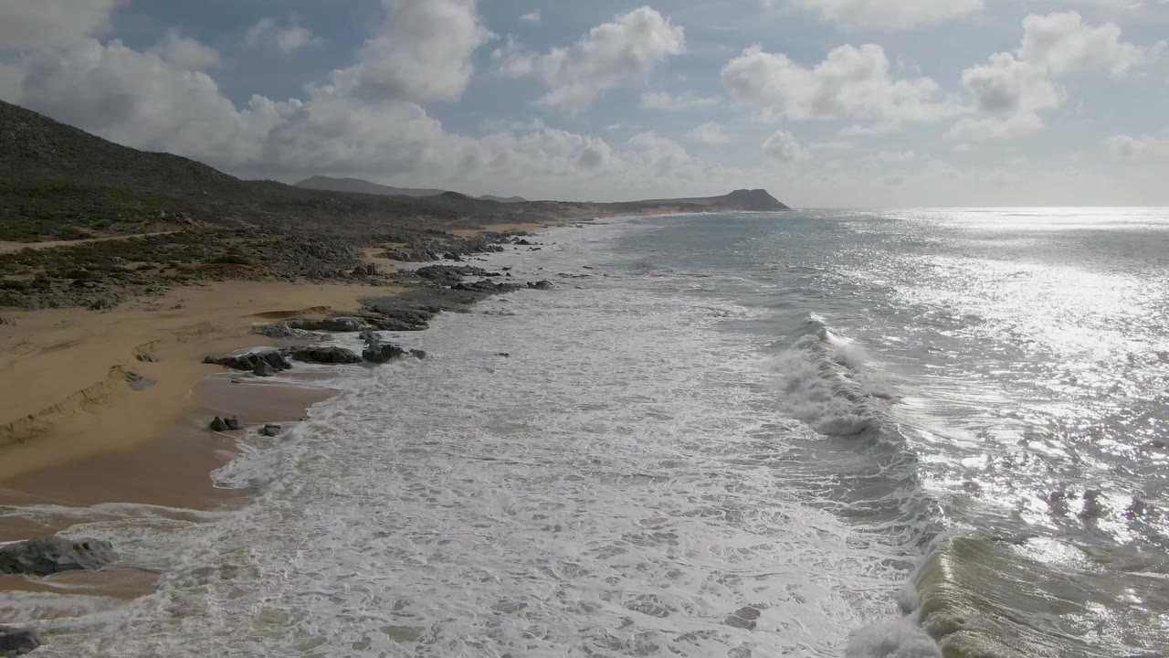 Aerial view at Punta Gorda, Baja California Sur, where foamy waves push toward a sandy beach with dark rocky outcrops, low coastal hills, bright water glare, and no visible people.