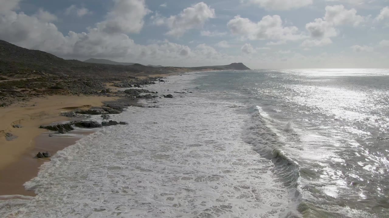At Punta Gorda, Baja California Sur, foamy surf covers more of the foreground water while a small sandy curve and scattered black rocks sit along the left shoreline with no visible people.