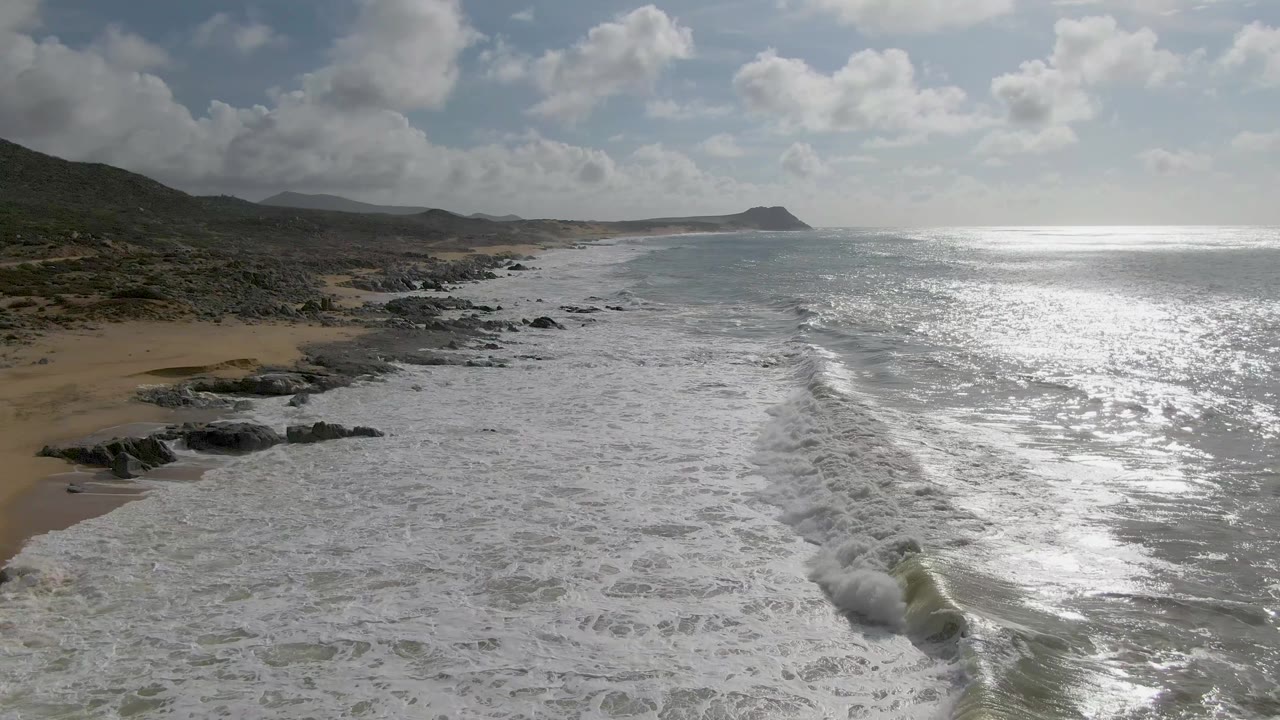 A breaking wave line angles through the foreground at Punta Gorda, Baja California Sur, with rocky beach on the left, low hills behind it, a bright ocean reflection, and no visible people.