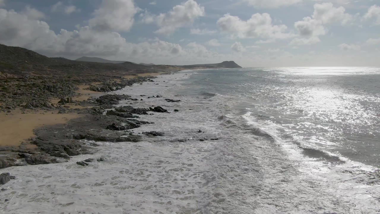 Elevated view of Punta Gordo East Cape shoreline with white waves rolling over dark rocks beside sandy beach, distant hills, bright ocean glare, and no visible people.
