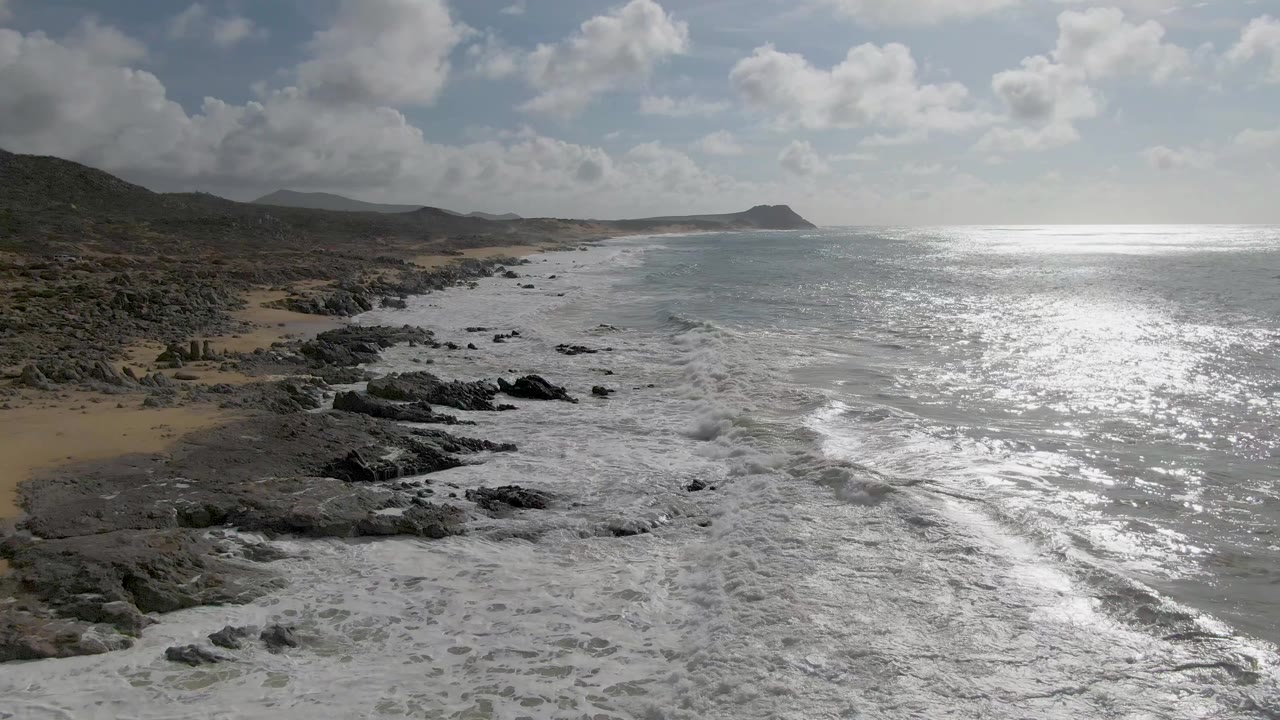 Punta Gordo East Cape surf washes farther across the rocky shoreline, with sand at the left edge, silver-blue water to the right, coastal hills beyond, and no visible people.