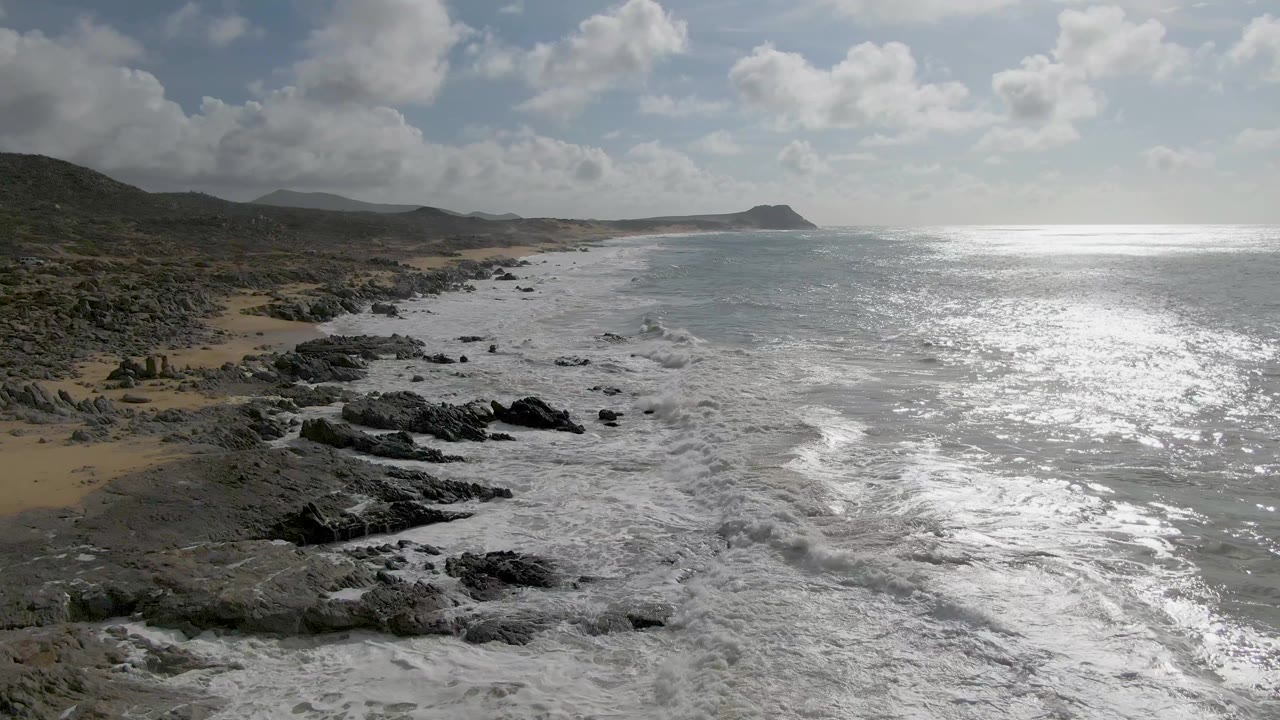 White water breaks over clustered dark rocks on the Punta Gordo East Cape coast, with a narrow sandy strip, shining sea surface, distant headland, and no visible people.