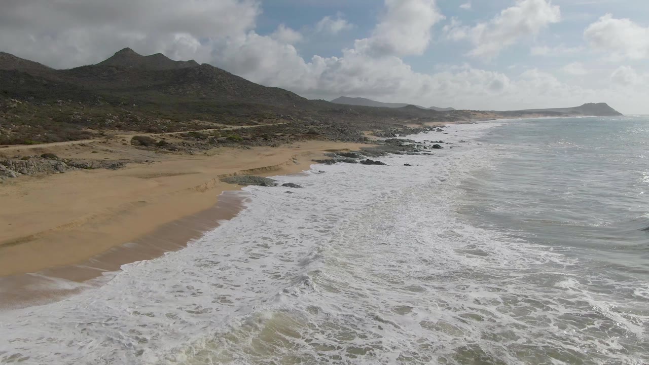 Aerial view of surf washing onto a sandy beach with mountains behind the shoreline.