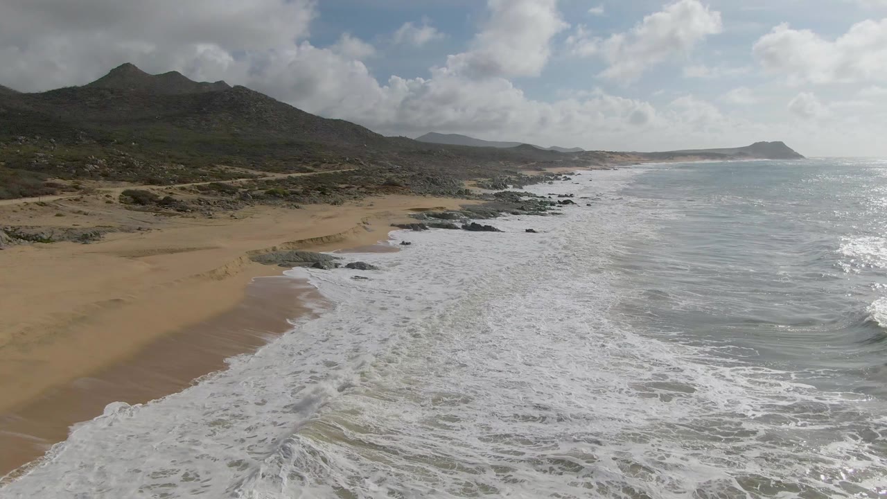At 0:01, white foam covers more of the nearshore water at Punta Gorda, with the sandy beach on the left, rocky points along the coast, distant mountains, and no visible people.