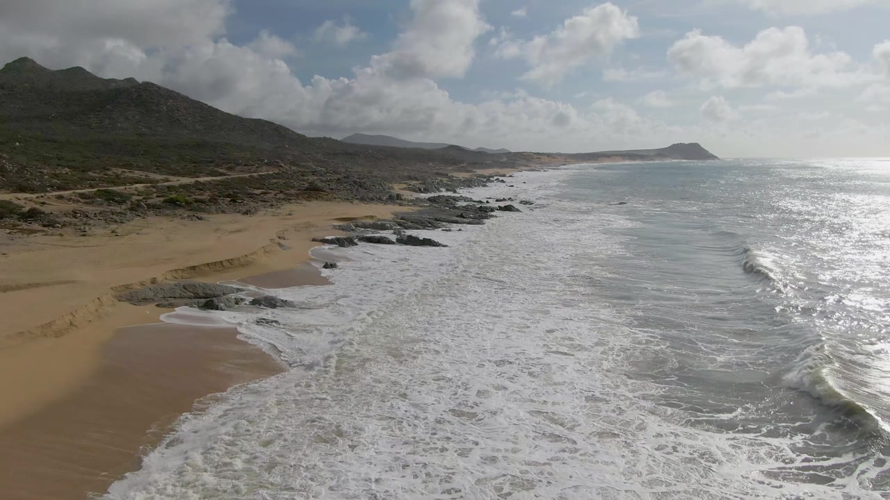 At 0:03, the view looks farther along the Punta Gorda shoreline, with foam running parallel to the beach, rocky ledges near the water, bright sea surface, mountains, and no visible people.