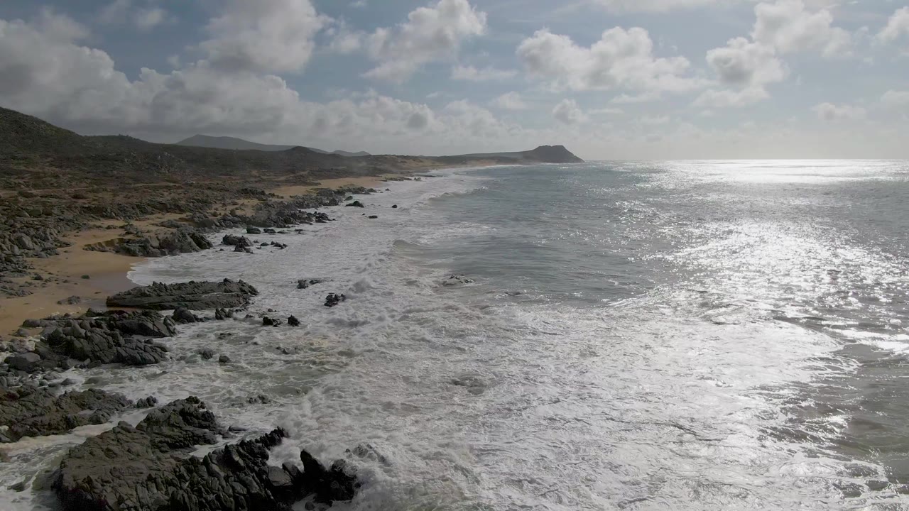 Wide elevated view at Punta Gorda, Baja California Sur, with white surf covering rocky outcrops, a sandy beach on the left, low hills inland, bright ocean glare, and no visible people.