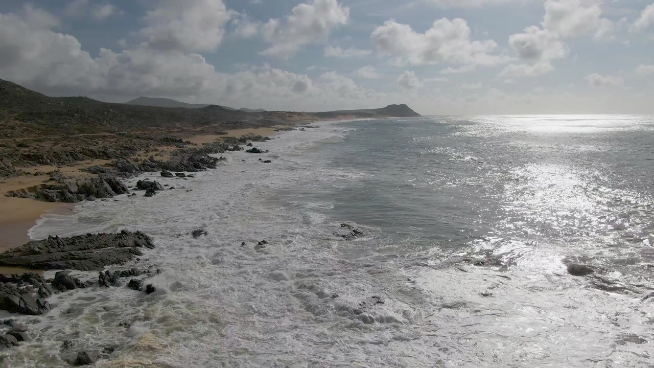 Punta Gorda shoreline appears with broken rocks in the foreground, sandy pockets along the beach, bright glare across the water to the right, inland hills, clouds, and no visible people.