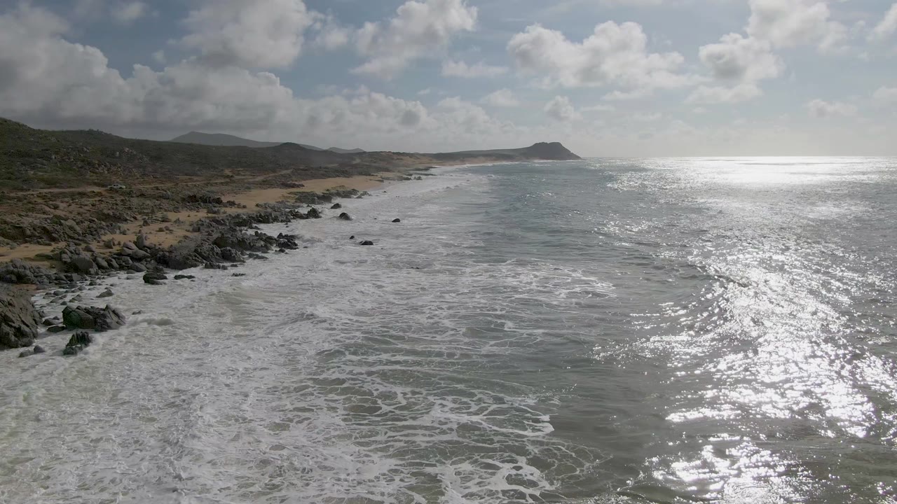 Aerial view of Punta Gorda East Cape shoreline with white surf spreading over gray-green water, dark rocks along tan sand, a distant headland, broken clouds, and no visible people.