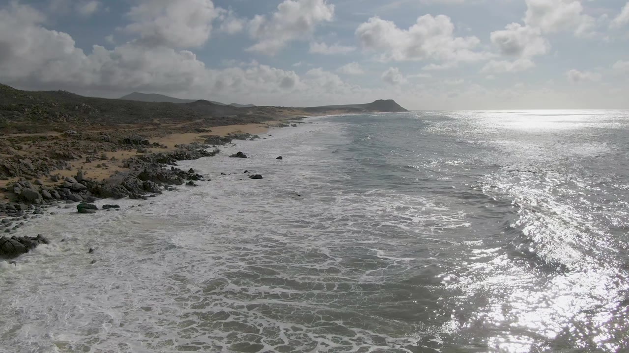 Aerial frame over Punta Gorda East Cape with breaking waves closer to the rocky shoreline, tan beach curving along the left, bright reflected light on the water, and no visible people.