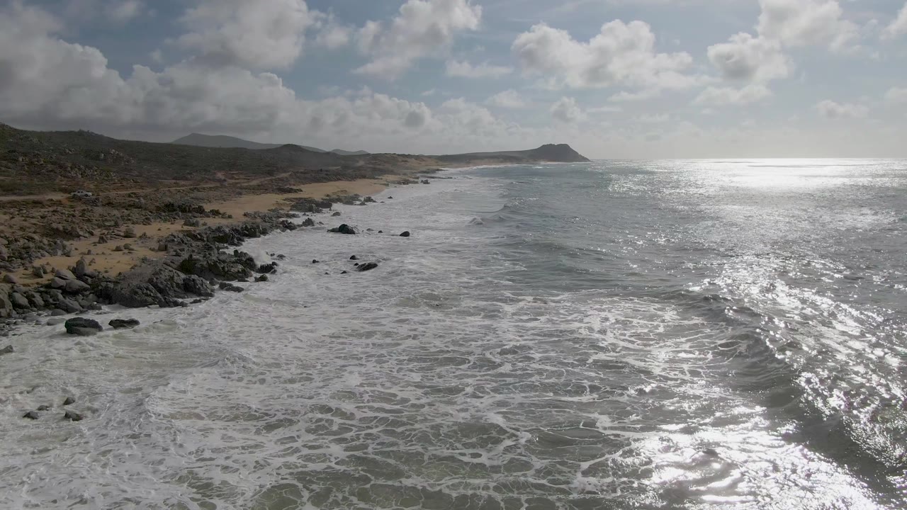 Aerial view at Punta Gorda East Cape showing wider foamy water in the foreground, scattered black rocks near shore, tan sand, low green-brown hills, a far headland, and no visible people.