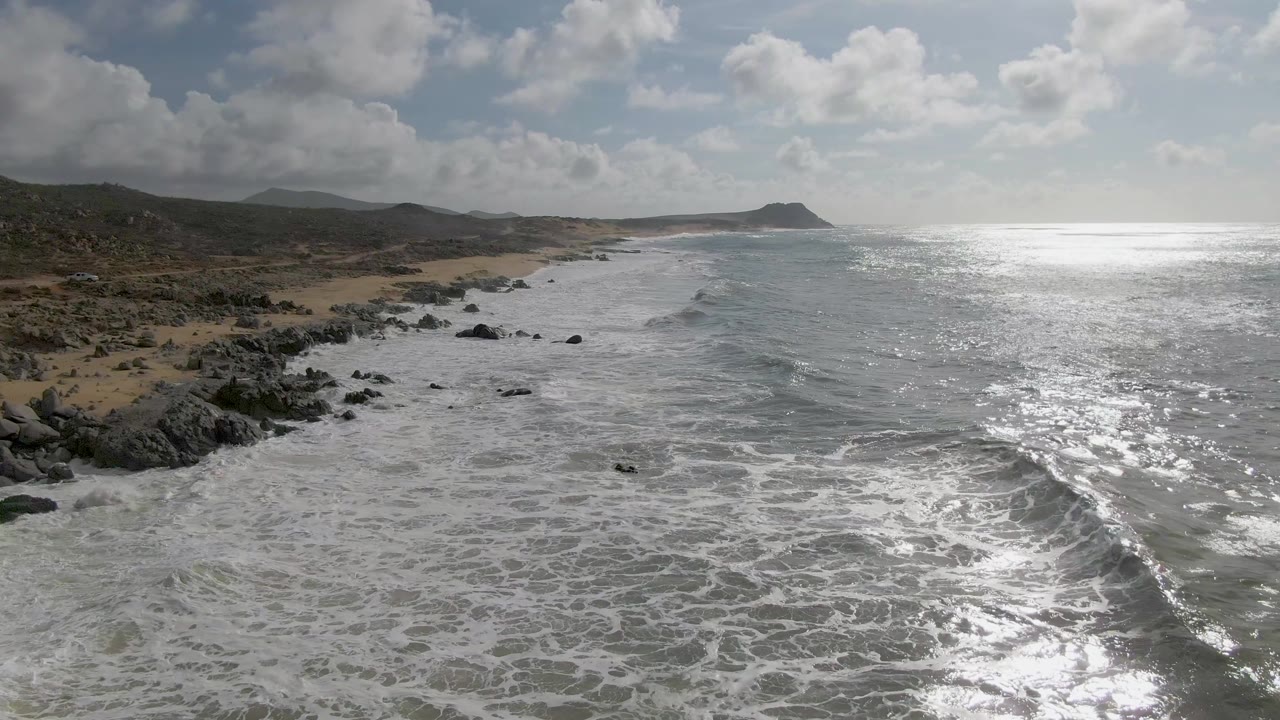Aerial frame from Punta Gorda East Cape with churning white surf below, a rocky and sandy shoreline to the left, bright glare across the right side of the ocean, and no visible people.