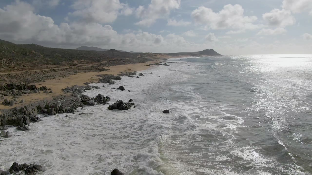 Punta Gorda coast seen from above with a broad band of foamy surf in the foreground, beach and rocky outcrops along the left, bright water to the right, and no visible people.