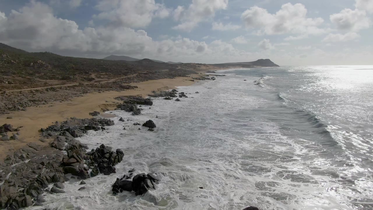 Aerial view at Punta Gorda, Baja California Sur, where white surf wraps around dark shoreline rocks beside tan sand, with no visible people.