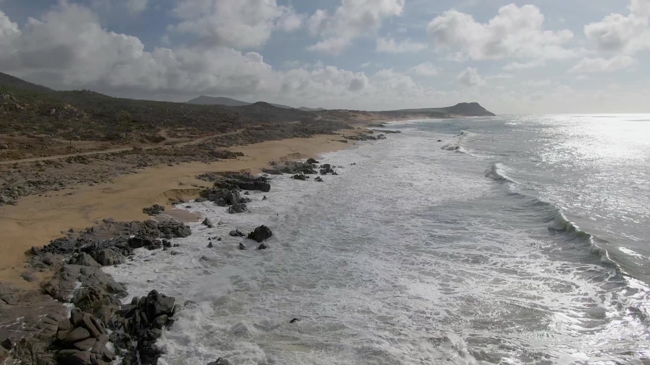 Aerial frame from Punta Gorda showing foamy waves breaking along a rocky sandy shore, bright reflected light on the ocean, and no visible people.
