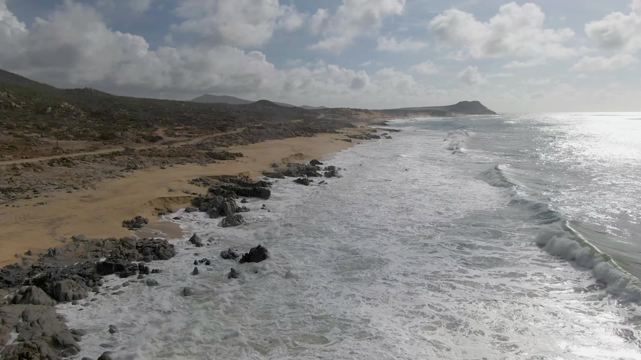 At Punta Gorda, dark rock outcrops divide white surf from the tan beach while a low hill and distant headland sit under broken clouds, with no visible people.