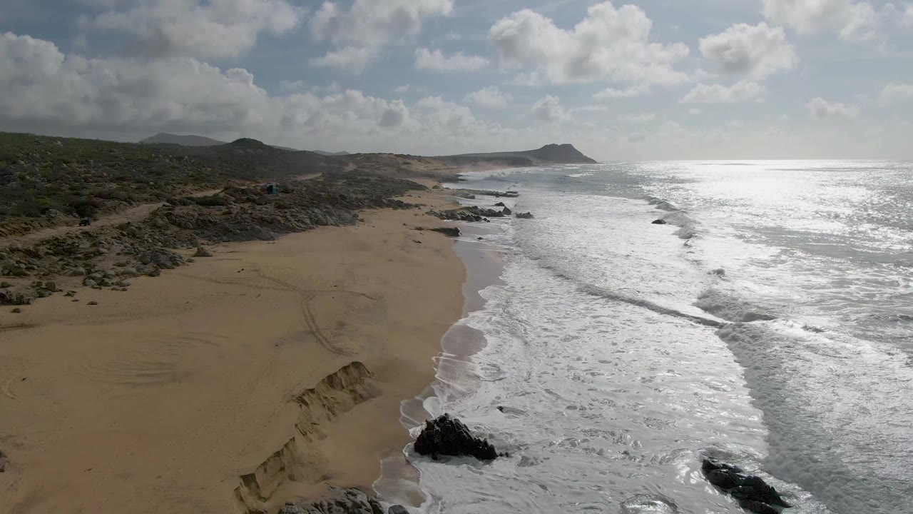 Aerial view at Punta Gordo, Baja California Sur, showing white surf washing toward a sandy beach with rocky outcrops, scrubby hills, bright ocean glare, and no visible people.