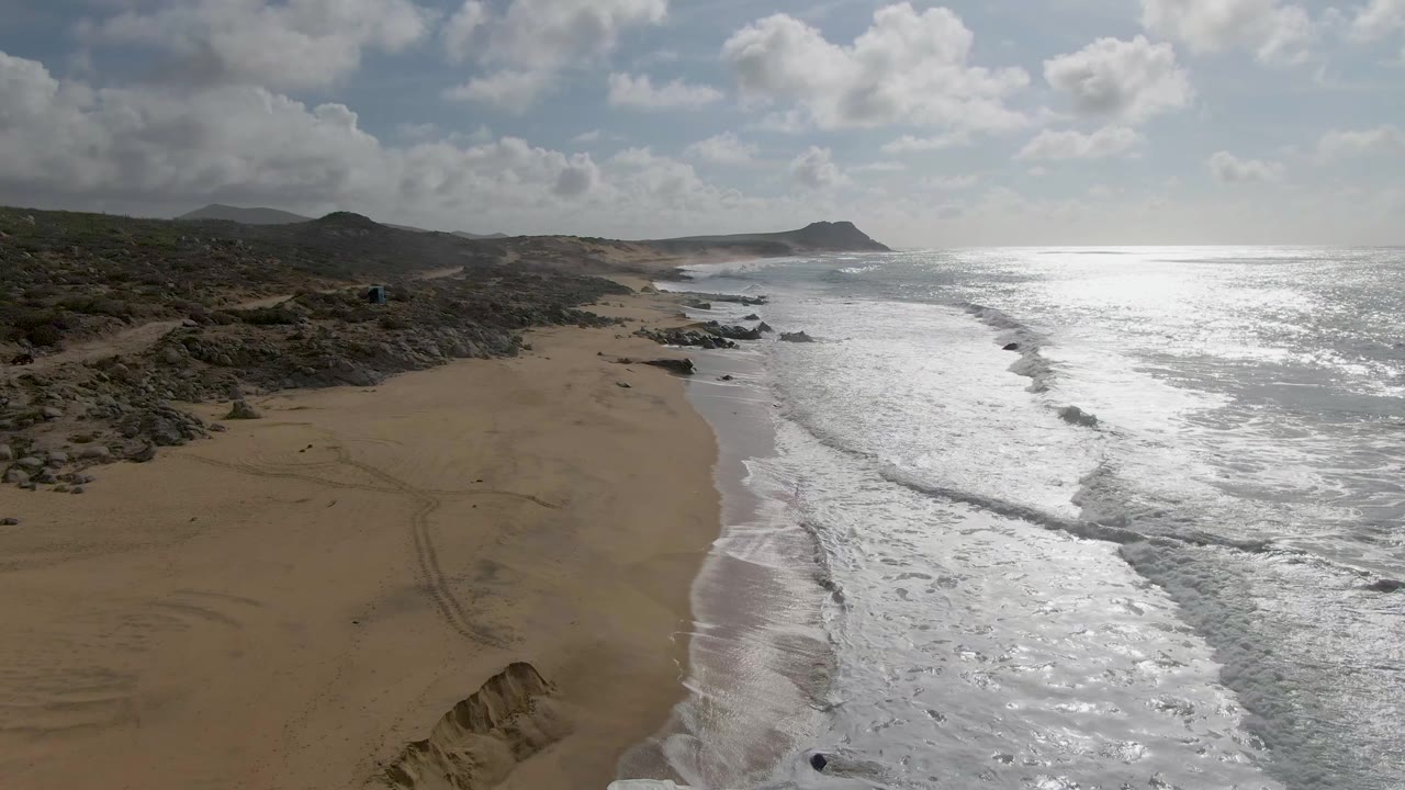 Aerial frame at Punta Gordo, Baja California Sur, with a broad band of foam along the beach, darker rocks near the waterline, scrub-covered hills inland, and no visible people.
