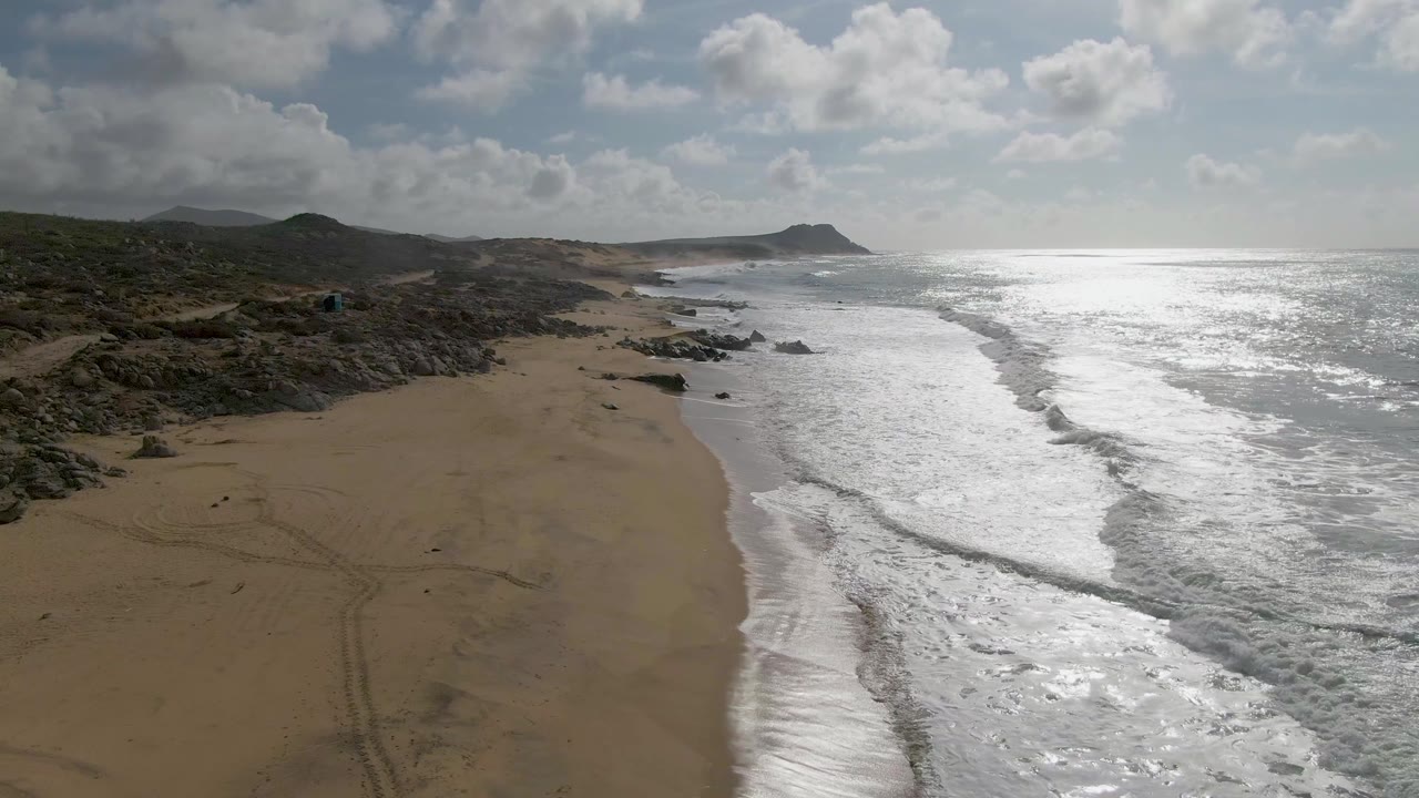 Aerial view at Punta Gordo, Baja California Sur, showing surf spread across the right side of the frame, an empty sandy beach to the left, scattered rocks near shore, and no visible people.