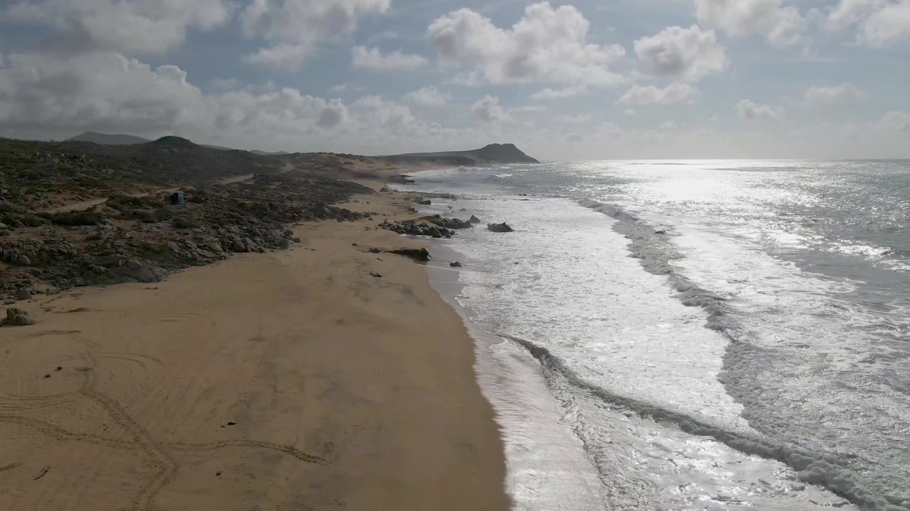 Aerial frame at Punta Gordo, Baja California Sur, with wet and dry sand divided by a bright surf edge, rocky terrain inland, ocean extending to the horizon, and no visible people.