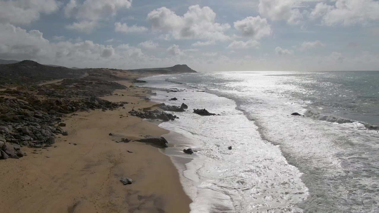 Aerial frame from Punta Gorda East Cape in Baja California Sur, Mexico, with white surf pushing along the sandy shoreline, dark rocks in the midground, reflective ocean to the right, and no visible people.