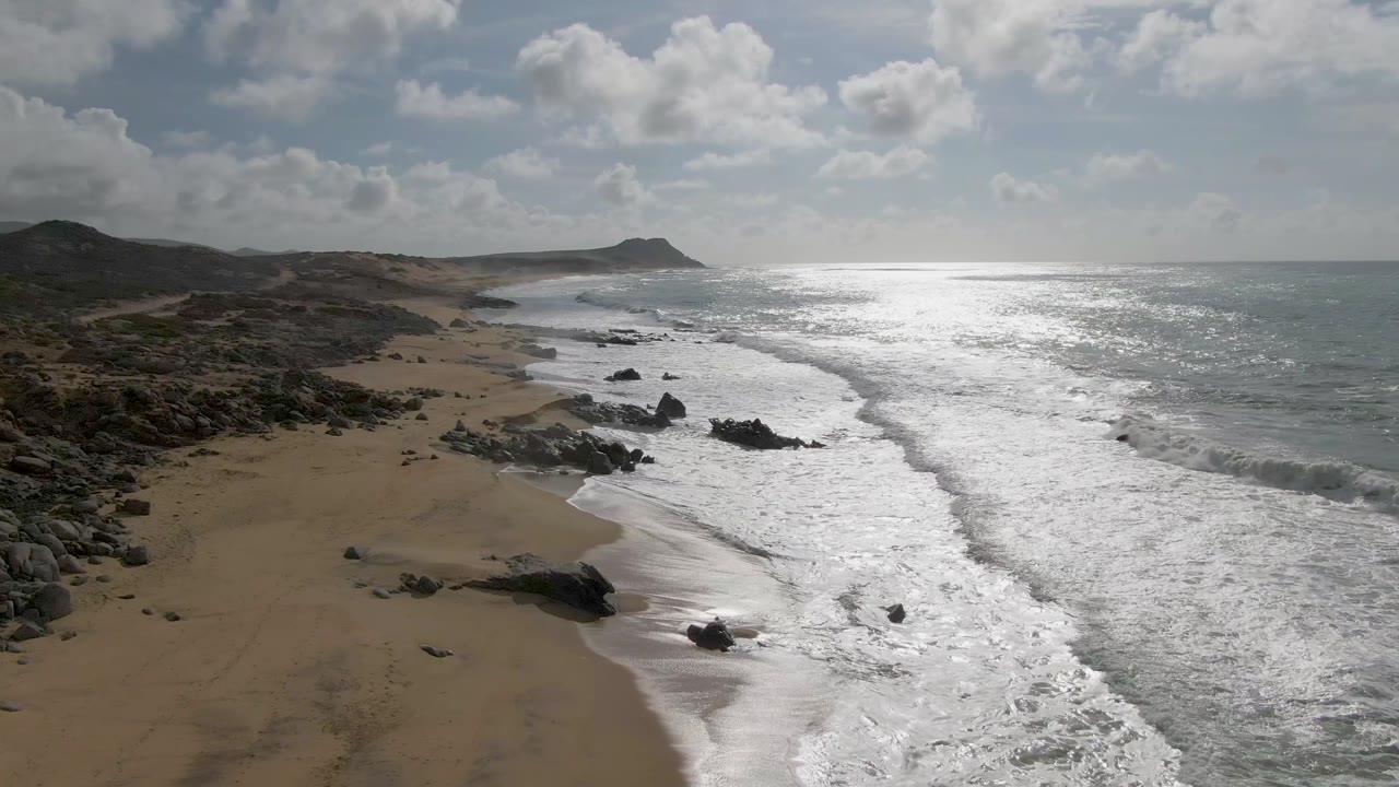 Punta Gorda East Cape coastal view in Baja California Sur, Mexico, with surf curling around rocks on the sand, a broad band of ocean glare, scattered clouds, low hills, and no visible people.
