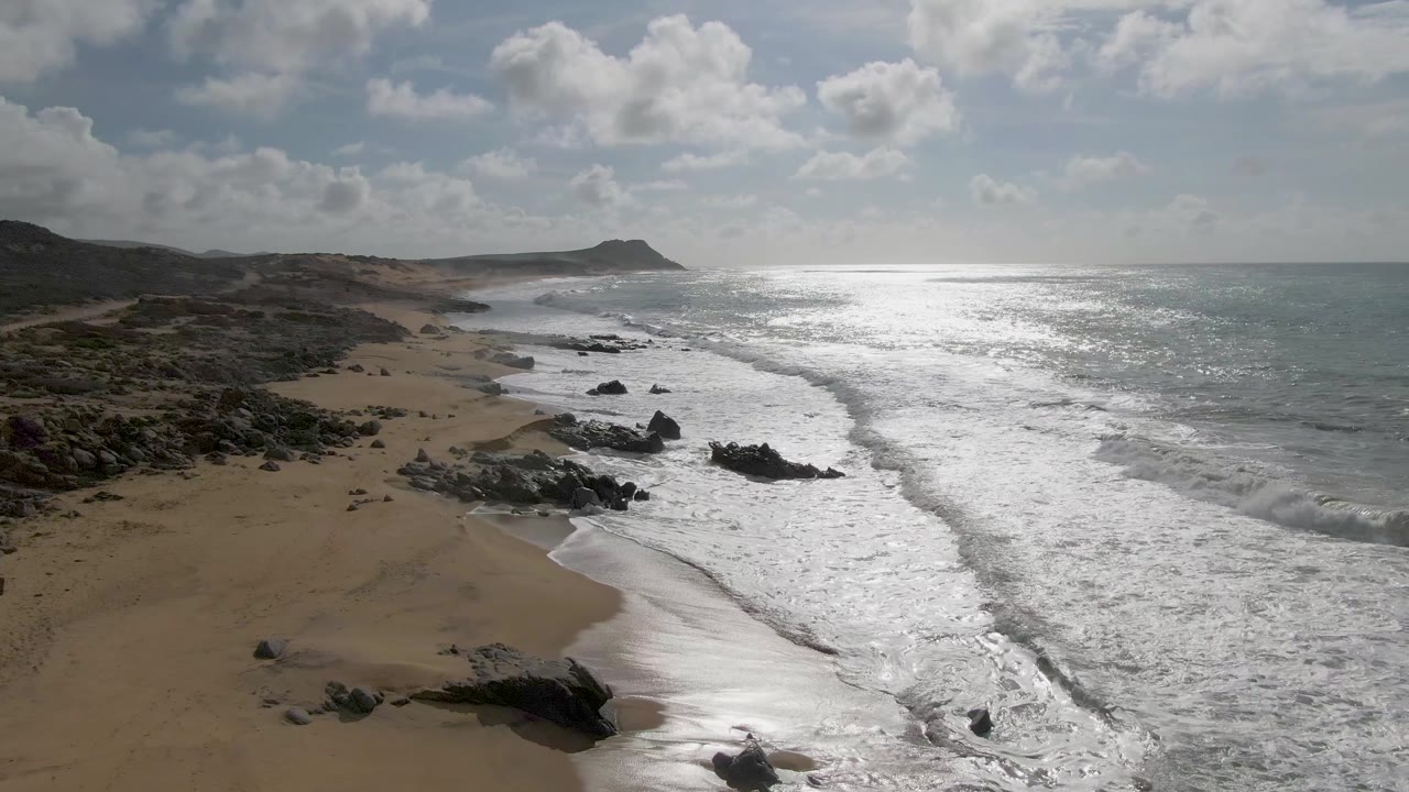 Wide aerial frame at Punta Gorda East Cape in Baja California Sur, Mexico, looking along the coastline where foamy waves meet sand and rocks beneath a cloudy sky, with no visible people.