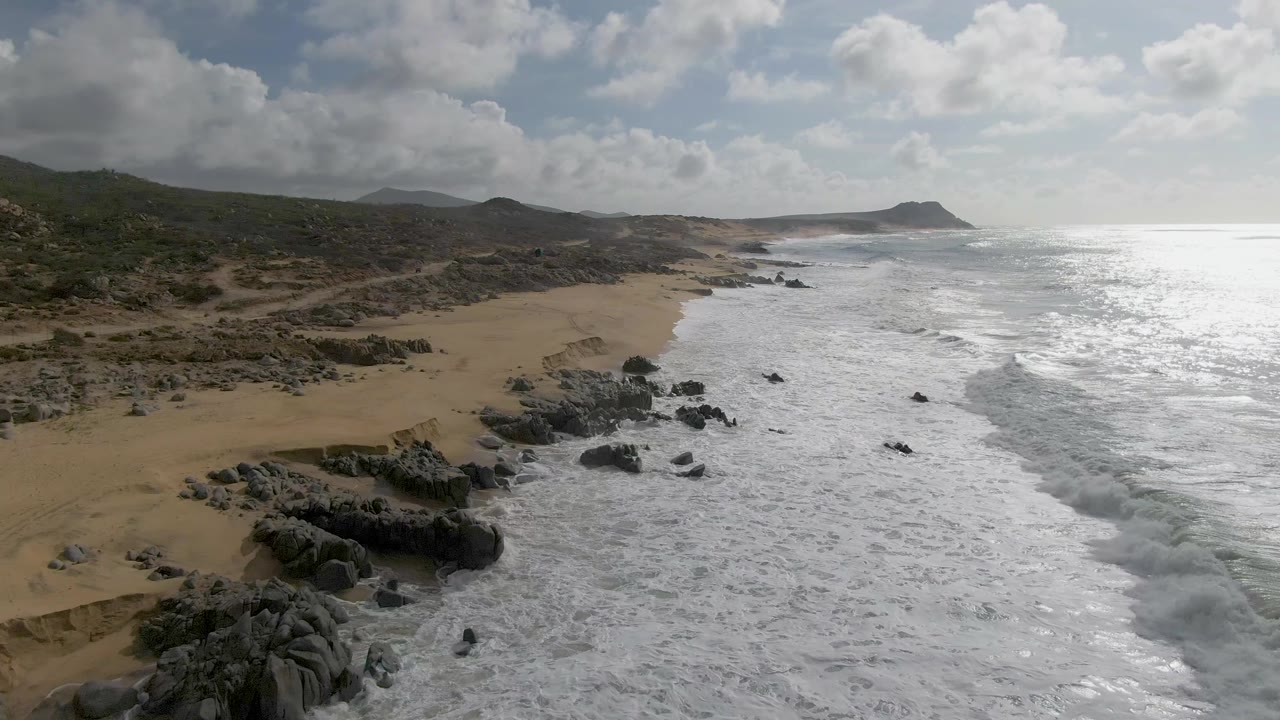At Punta Gorda, Baja California Sur, an aerial view looks along a sandy coast where white surf breaks around dark rocks, with scrub hills inland and no visible people.