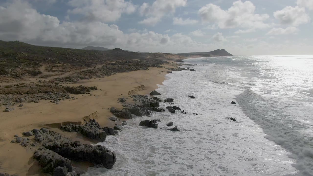 At Punta Gorda, Baja California Sur, whitewater spreads farther across the nearshore rocks while the tan beach curves beneath low green-brown hills, with no visible people.