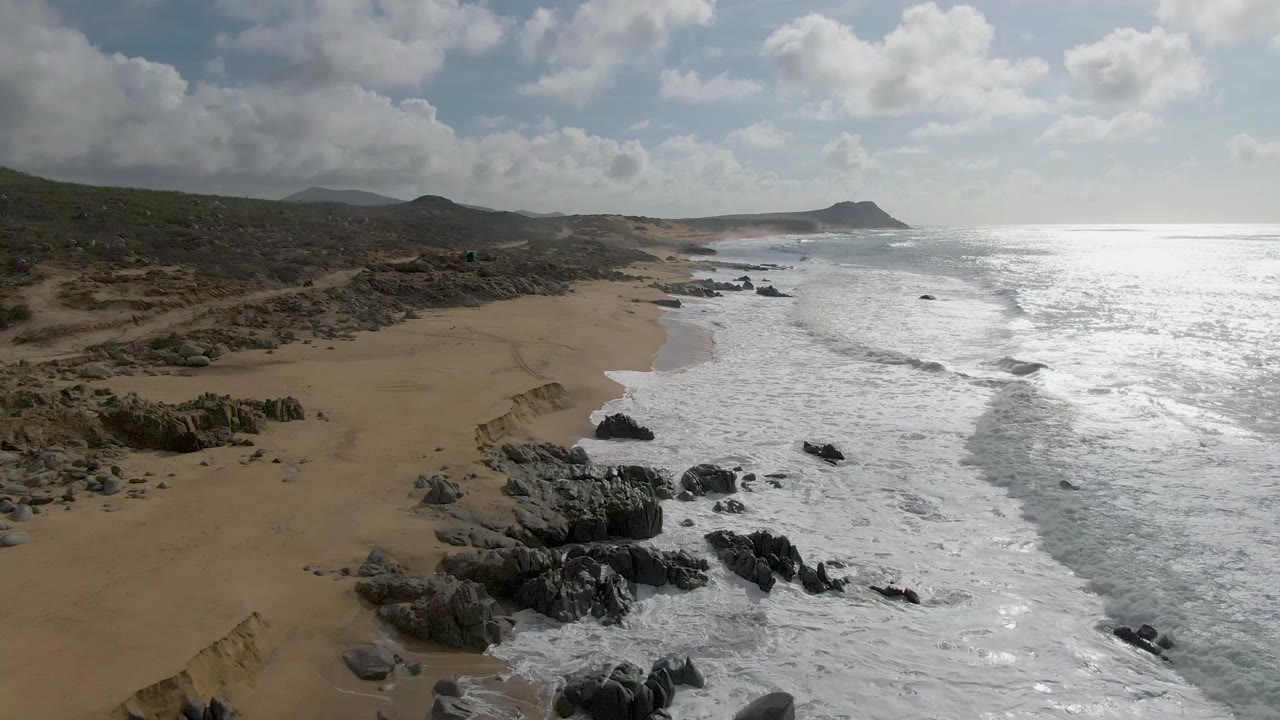 Foamy waves run beside a broad empty sandy beach with rocks below.