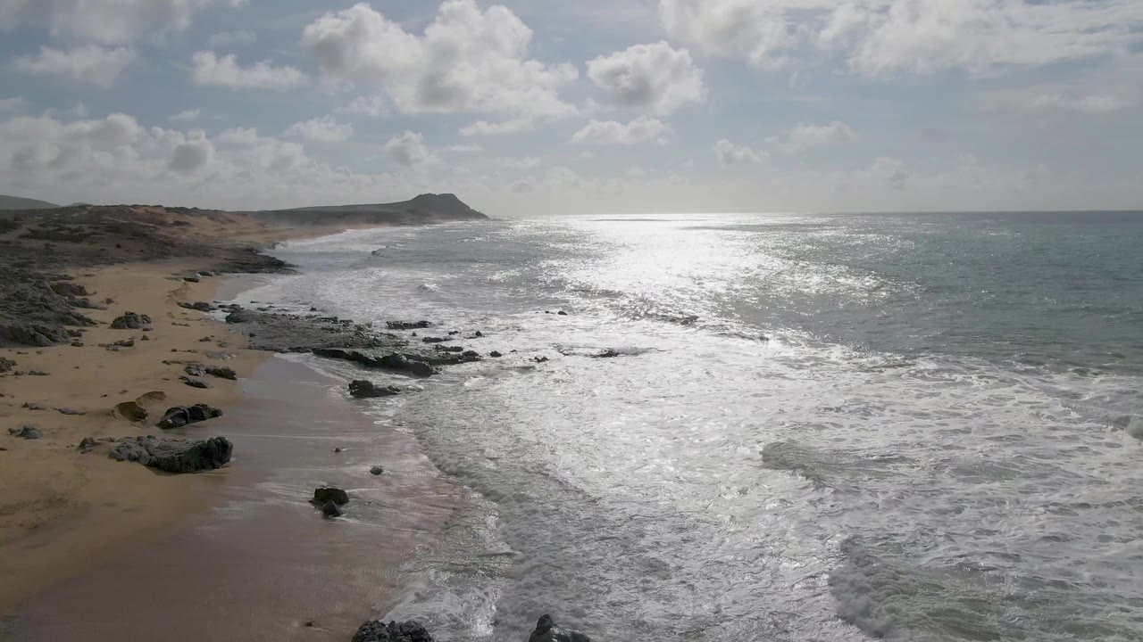 Aerial view at Punta Gorda, Baja California Sur, with white waves spreading over dark shoreline rocks, wet sand on the left, bright ocean glare to the right, and no visible people.