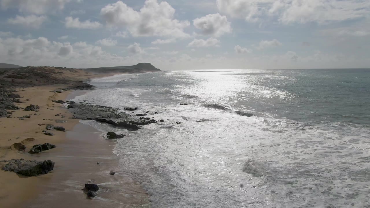 At 0:01, the Punta Gorda shoreline remains angled through frame as foamy water covers more of the rocks, with a sandy beach at left, sunlit sea at right, and no visible people.