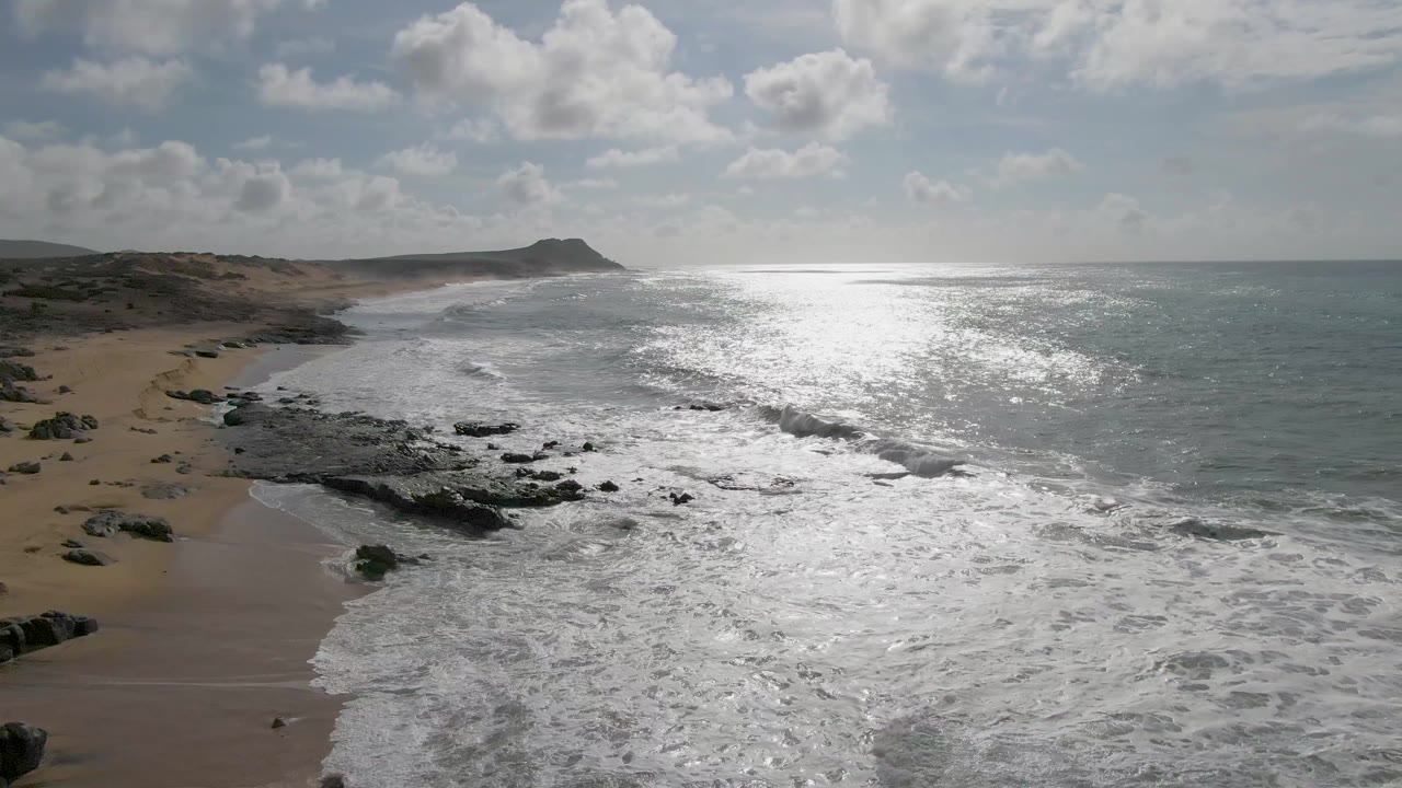 At 0:02, white surf advances along the rocky Punta Gorda shore, with tan beach and low coastal land to the left, bright reflected light on the ocean, scattered clouds, and no visible people.