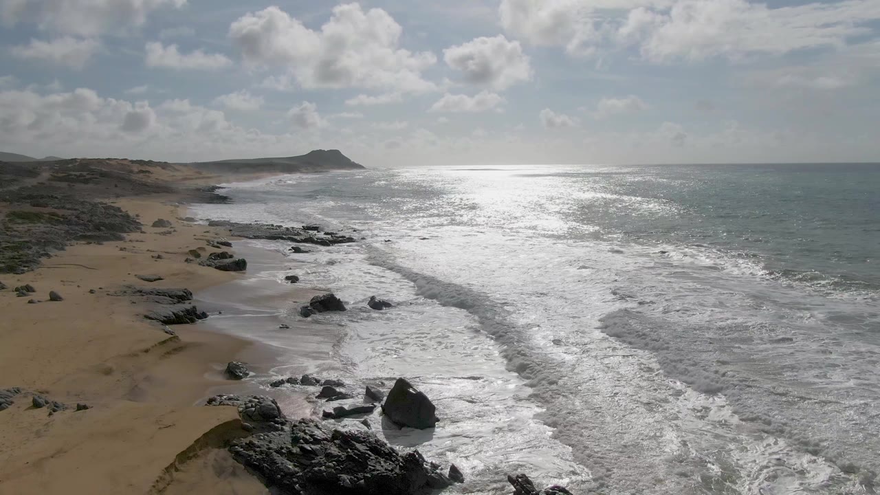 Aerial coastal frame from Punta Gorda with a large dark rock near the wash, foamy surf moving diagonally across the beach, bright water glare, and no visible people.