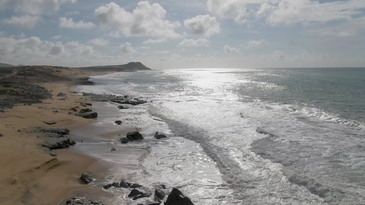 Punta Gorda beach from an elevated angle, with scattered rocks in the lower frame, foam lines crossing the wet sand, open ocean glare, and no visible people.