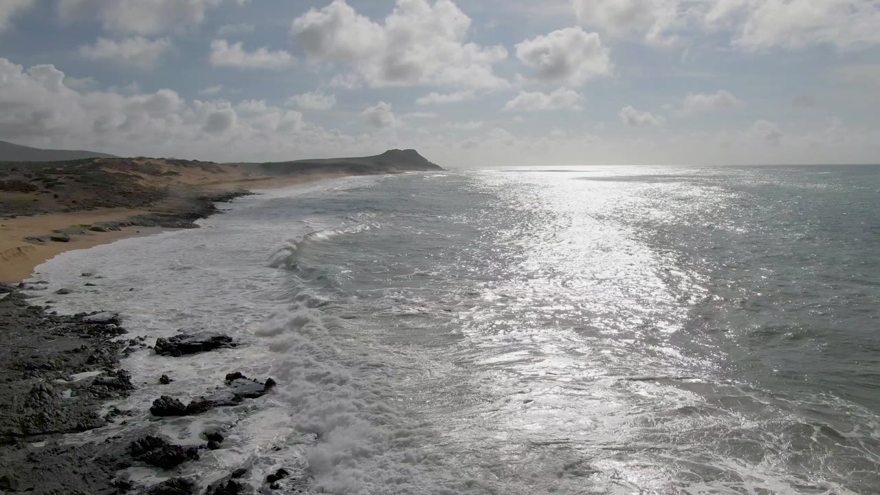 Aerial view at Punta Gorda, East Cape, Baja California Sur, with white surf spreading over dark foreground rocks, sandy shoreline to the left, bright ocean glare to the right, and no visible people.