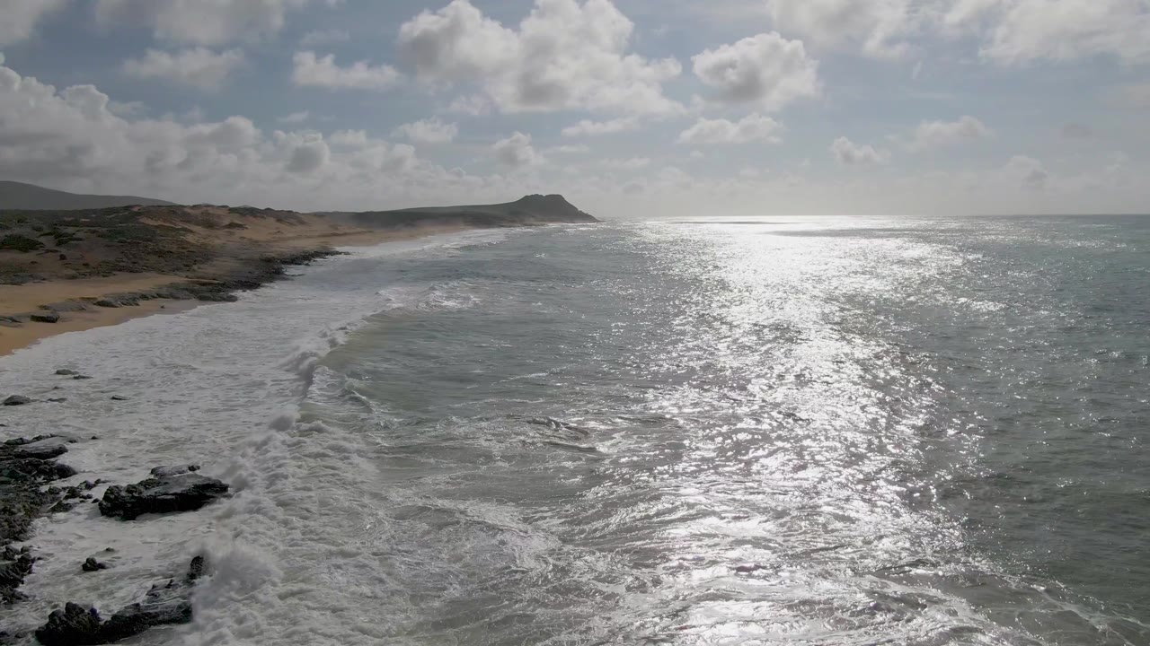 At 0:01, the Punta Gorda coast remains in a wide aerial composition as foam pulls along the rocky edge, the sandy beach curves into the distance, and no visible people appear in frame.