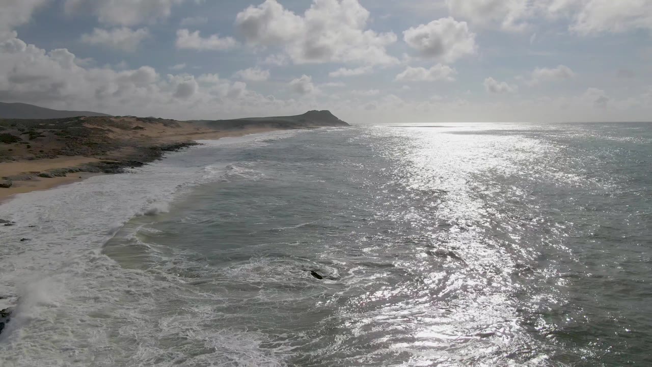 At Punta Gorda, East Cape, white surf covers more of the near shoreline while the ocean surface on the right flashes with sunlight under scattered clouds, with no visible people.