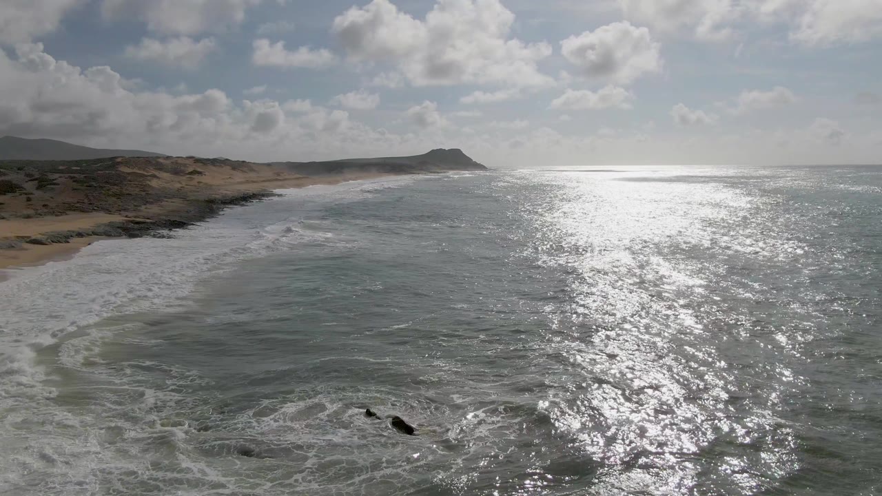 Breaking waves fan across rocks and sand under a partly cloudy sky.