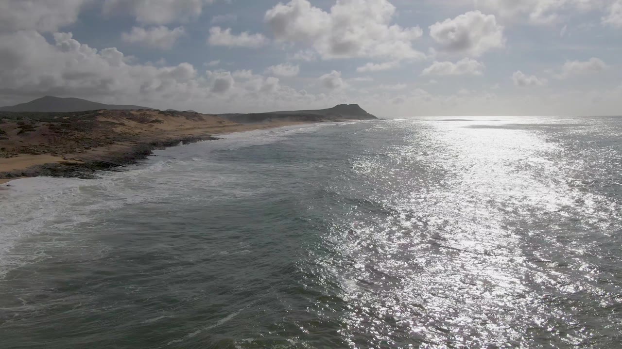 Aerial-looking view at Punta Gorda, Baja California Sur, with green-blue ocean in the foreground, white surf along a sandy and rocky shore, distant hills, bright sun reflection on the water, and no visible people.