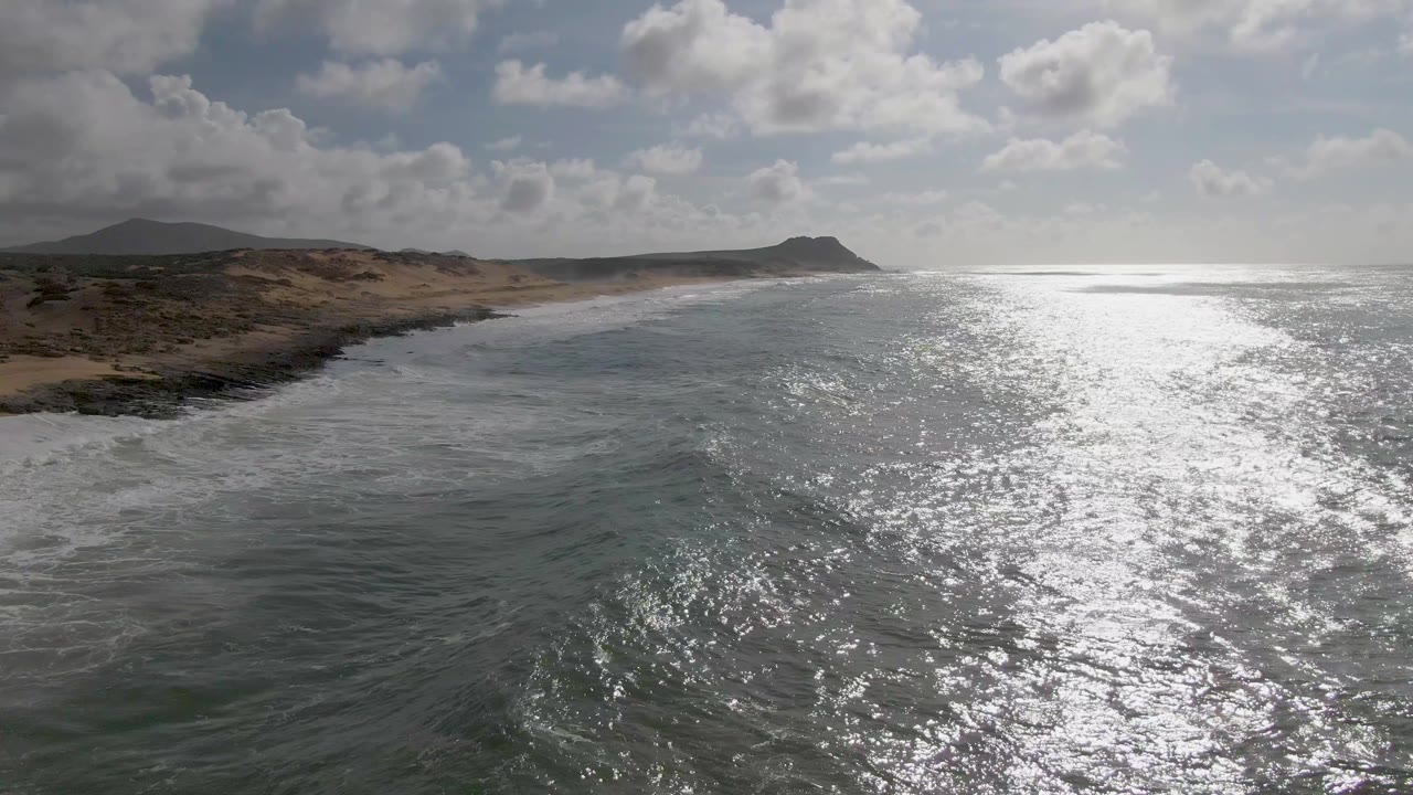At 0:01, Punta Gorda coastline remains at left while the sunlit water fills the center, with white wave foam tracing the shore and no visible people.