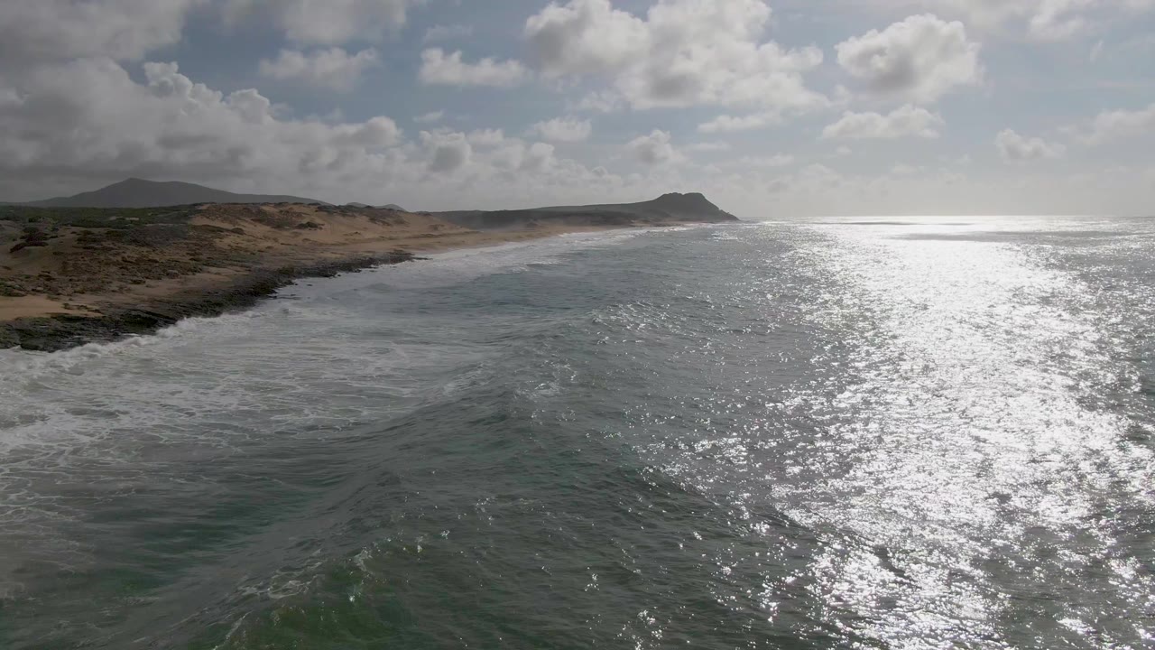 At 0:02, the Punta Gorda shore sits left of center with waves breaking into white foam, open water and glare to the right, low hills beyond, and no visible people.