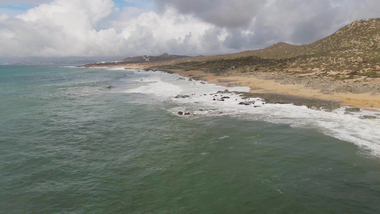 Aerial view of waves reaching a sandy beach with rocky outcrops and hills beyond.